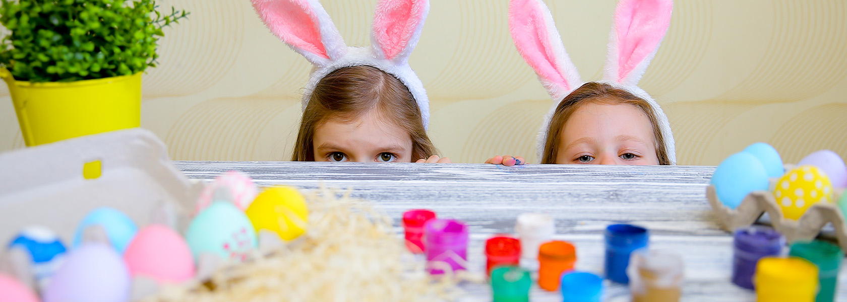 Zwei Kinder mit Hasen-Ohren auf dem Kopf schauen über die Tischkante auf einen Tisch mit bemalten Eiern und Malutensilien.